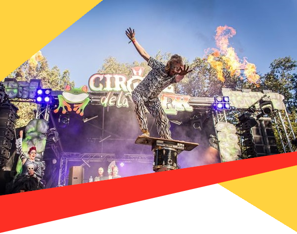 man balancing on board on precarious pile of objects in front of colourful stage